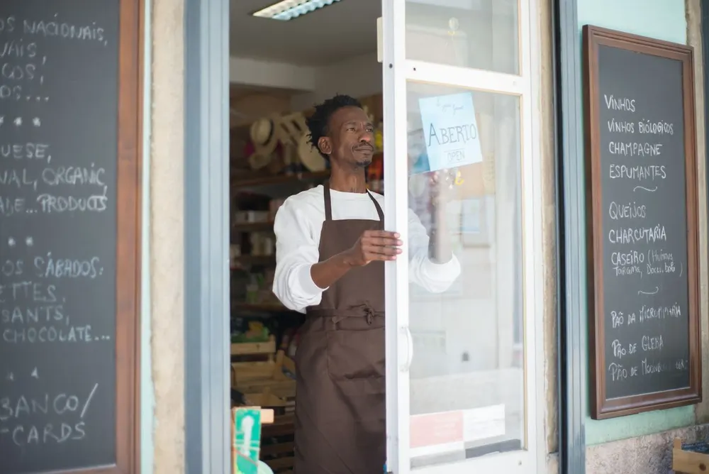 Comerciante abrindo a porta da própria loja, com a placa Aberto pendurada no vidro